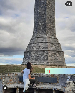 A white female with long brown hair sits looking at Hardy's monument. 